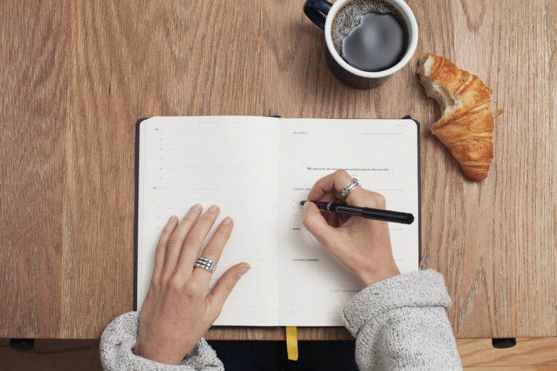 hands writing in a journal with cup of coffee and croissant